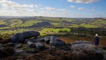 Curbar Edge View The landscape photograph captures Curbar Edge View in Derbyshire, United Kingdom, during the early afternoon in autumn. The scene showcases the rugged natural beauty of the Peak District, with expansive rural fields, rolling hills, and patchwork farmland stretching into the distance. Large rock formations are prominent in the foreground, characteristic of Curbar Edge, while the lush countryside is dotted with scattered trees and hedgerows. A person stands near the edge, observing the landscape, which highlights the harmonious relationship between humans and nature in this part of the United Kingdom. The photograph provides a clear illustration of the rural scenery for which Derbyshire and the Peak District are renowned.